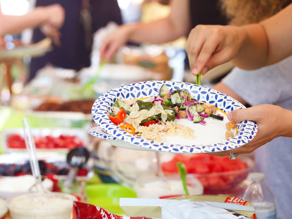 A person serves salad and pasta onto a paper plate at a summer BBQ picnic buffet, with various dishes and snacks visible on the table.