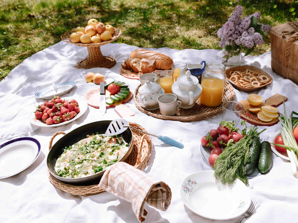 A summer BBQ picnic setup on a white blanket with various foods including bread, fruit, vegetables, tea, juice, and a skillet dish, all arranged outdoors in sunlight.