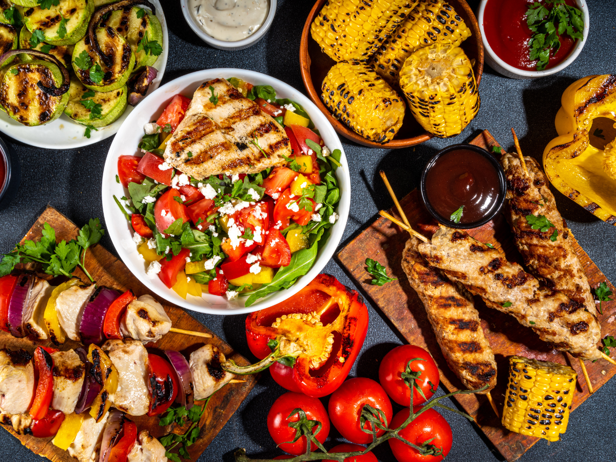 Grilled chicken salad, vegetable and meat skewers, corn on the cob, fresh tomatoes, and assorted sauces arranged on a table for a perfect summer BBQ picnic.