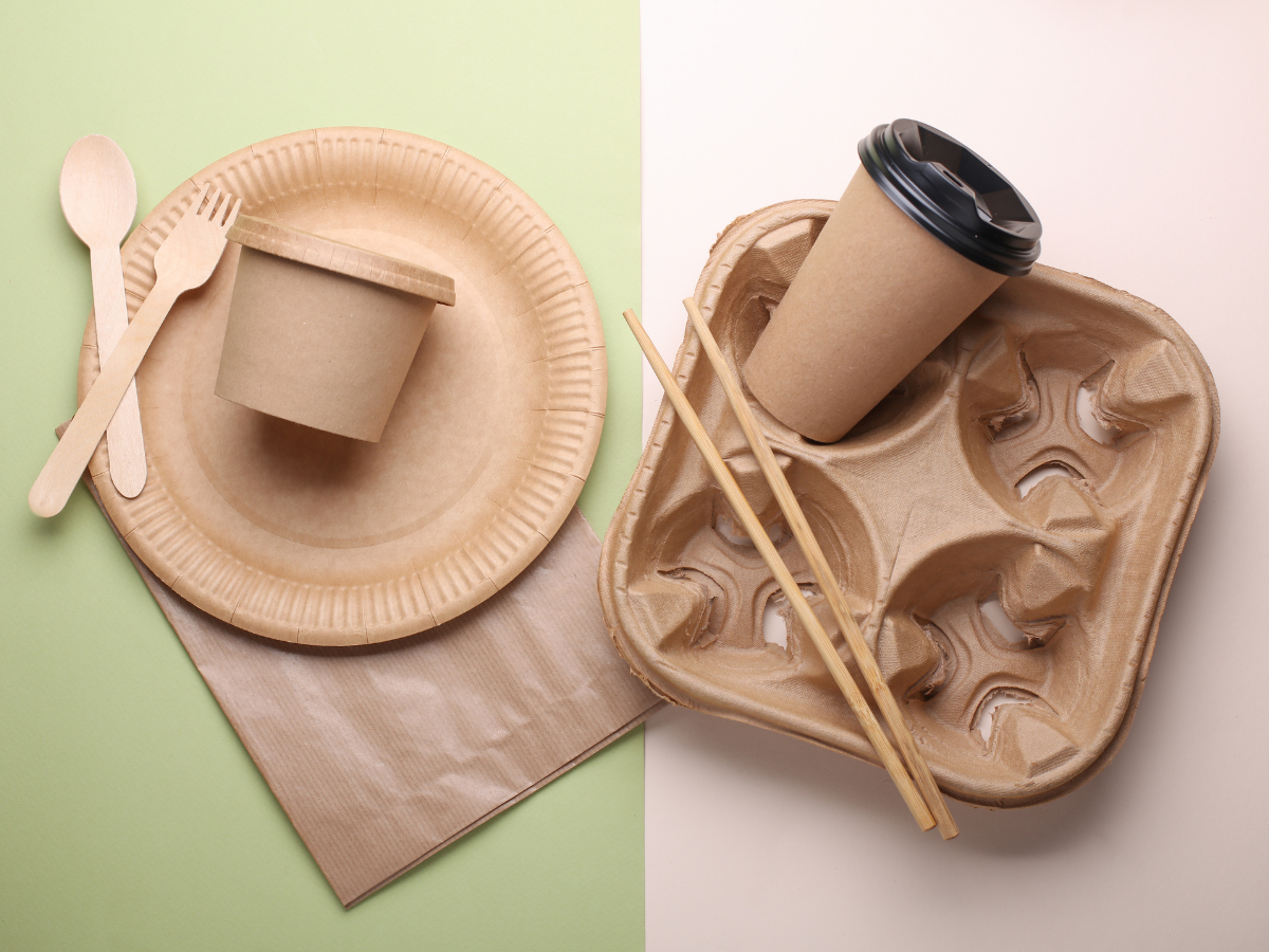 Eco-Friendly Packaging Ideas for Modern Catering 1 Assorted biodegradable tableware, featuring a paper plate, cup, wooden cutlery, chopsticks, and a cup holder arranged on a split green and beige background, showcases stylish eco-friendly packaging for your sustainable gatherings.