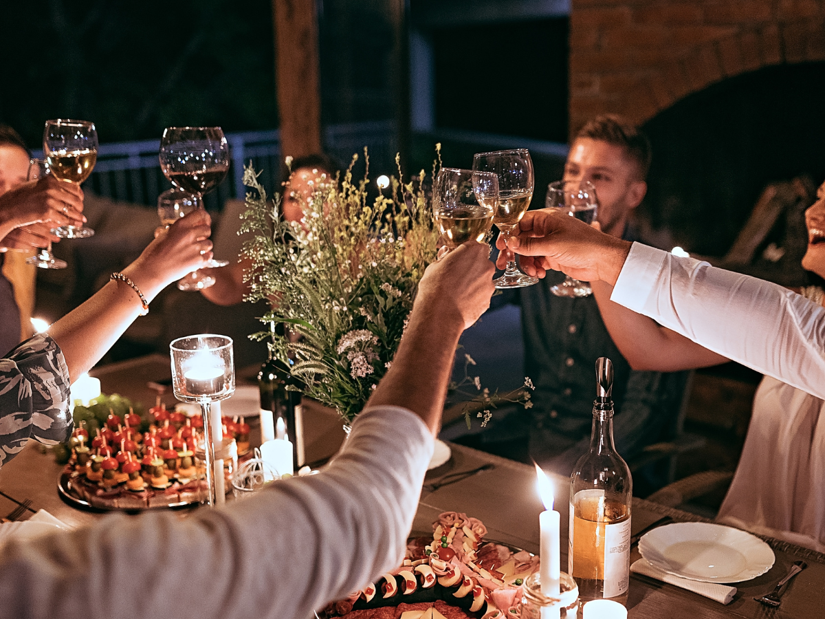 People seated around a candlelit table raise glasses in a toast. The table is set with flowers, food platters, wine, and candles—each detail thoughtfully arranged for the occasion. The scene appears warm and festive, capturing the joy and elegance of an expertly planned rehearsal dinner catering experience.