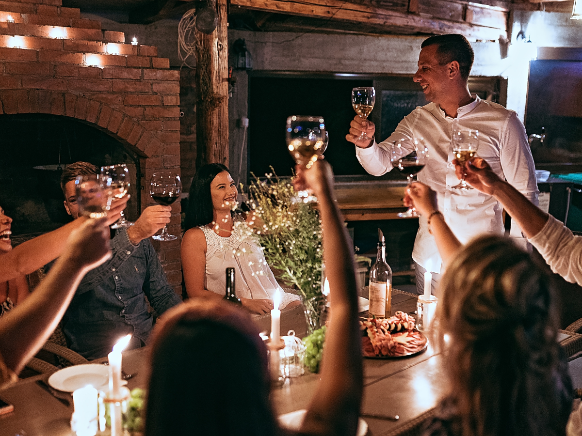Charming Rehearsal Dinner Catering in Metro Atlanta: Personalized Wedding Catering Solutions 3 A group of people sit around a candlelit table, raising glasses in a toast, with food and wine bottles in the center—a perfect scene for an unforgettable rehearsal dinner catering experience.