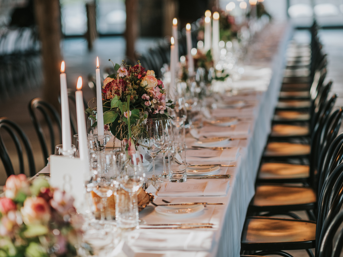 Charming Rehearsal Dinner Catering in Metro Atlanta: Personalized Wedding Catering Solutions 4 A long, elegantly set dining table with a white tablecloth, floral arrangements, lit candles, glassware, and plates showcases the perfect setting for a rehearsal dinner catering event in a spacious room with black chairs.