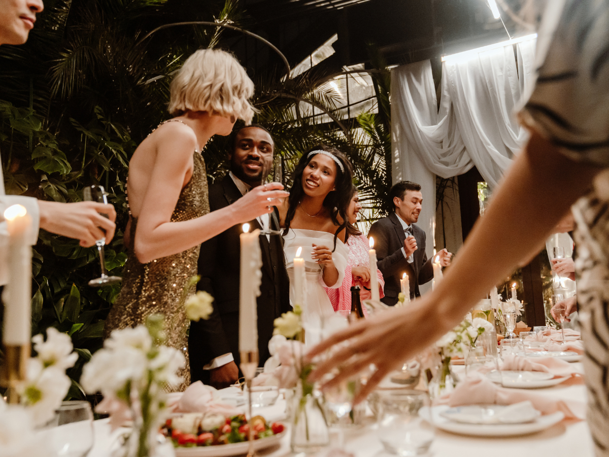 Charming Rehearsal Dinner Catering in Metro Atlanta: Personalized Wedding Catering Solutions 5 People dressed formally gather around a decorated dinner table with candles, flowers, and plates, engaging in conversation and celebration in an elegant, indoor setting enhanced by exquisite rehearsal dinner catering.