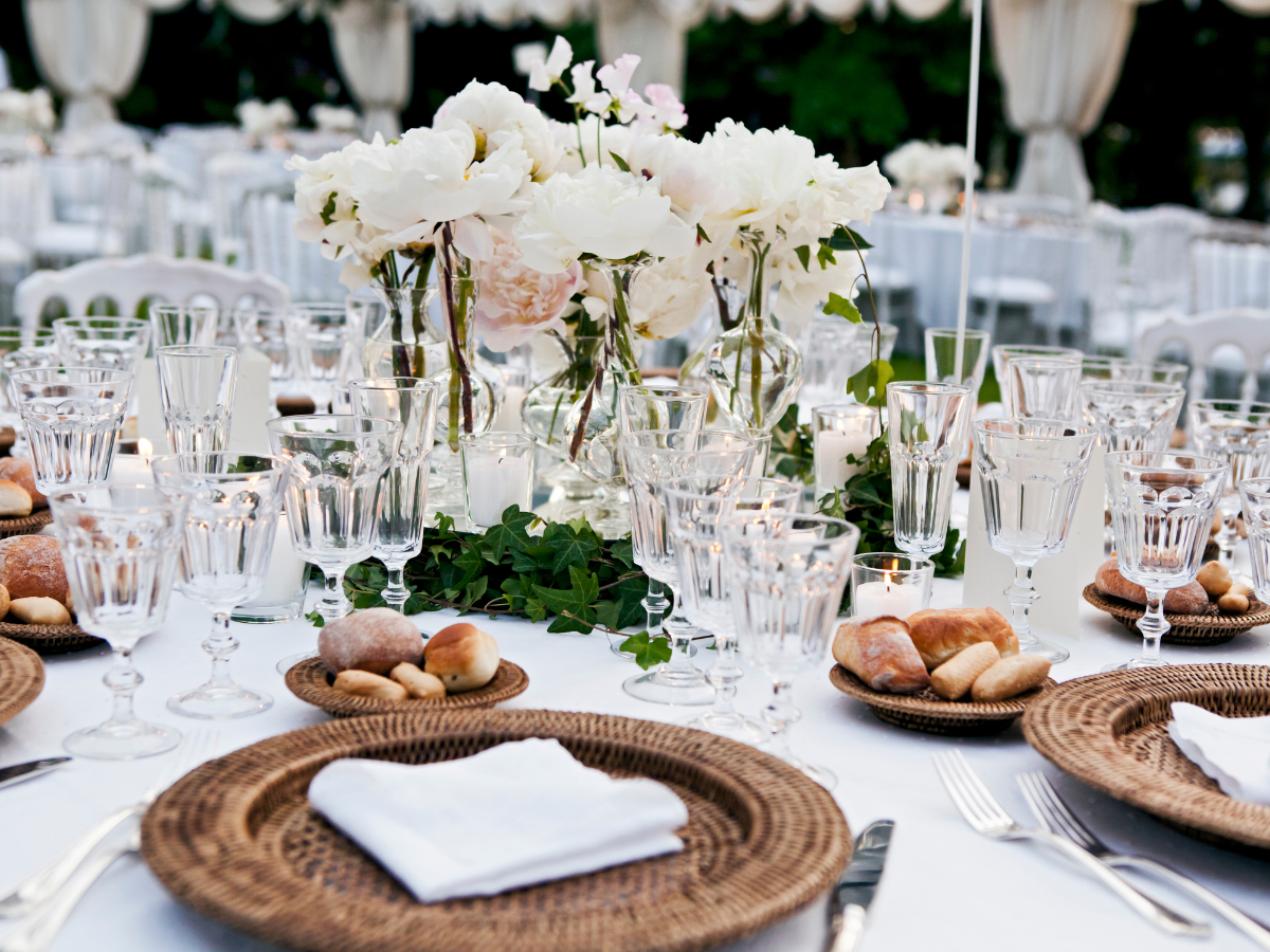 Charming Rehearsal Dinner Catering in Metro Atlanta: Personalized Wedding Catering Solutions 7 Elegant outdoor dining table set with woven placemats, white napkins, crystal glassware, bread rolls, and a floral centerpiece with white flowers and greenery—perfect for an intimate rehearsal dinner catering experience.