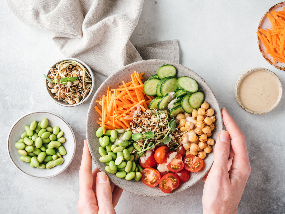 Hands holding a bowl with sliced cucumbers, cherry tomatoes, shredded carrots, chickpeas, edamame, and sprouts—perfect for a fresh vegetarian catering menu. Nearby are bowls of sprouts, edamame, shredded carrots, and a small bowl of dressing.