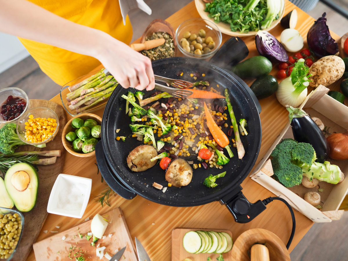 Person cooking assorted vegetables including carrots, corn, and potatoes on a large electric skillet, preparing a delicious vegetarian catering spread surrounded by fresh produce and cooking ingredients on a wooden table.