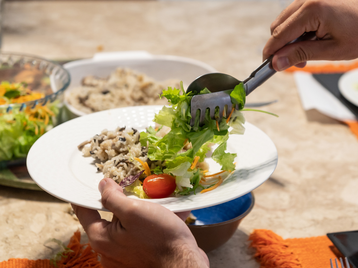 A person serves salad with greens, shredded carrots, and tomatoes onto a white plate, with rice and food in the background on a table, showcasing a colorful spread typical of vegetarian catering.