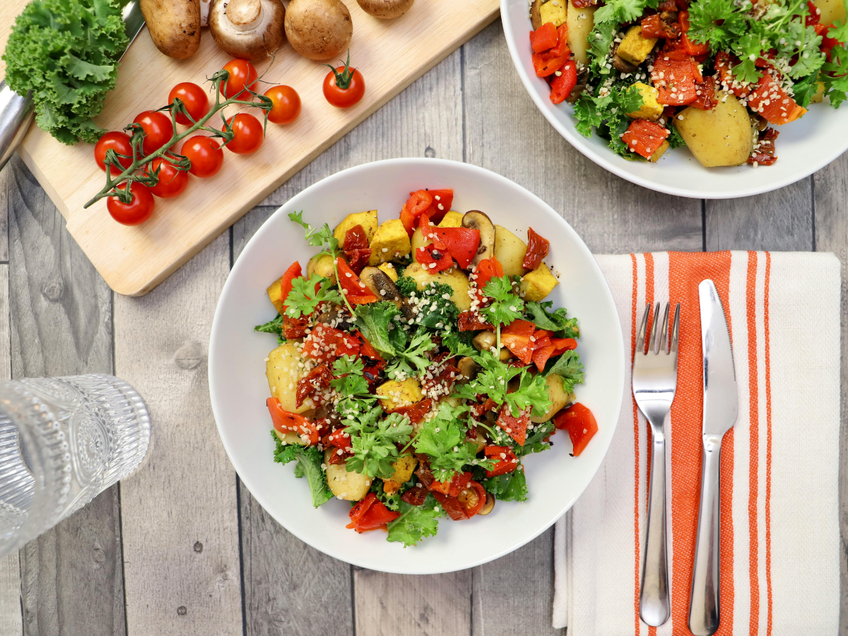 Bowl of roasted potatoes, cherry tomatoes, red peppers, greens, and sesame seeds on a table with a fork, knife, napkin, and glass of water—a vibrant display perfect for vegetarian catering.