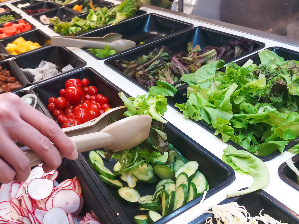 A person uses tongs to pick mixed greens from a salad bar featuring cherry tomatoes, cucumbers, leafy greens, radishes, and other fresh vegetables in black trays—an inviting display perfect for vegetarian catering.