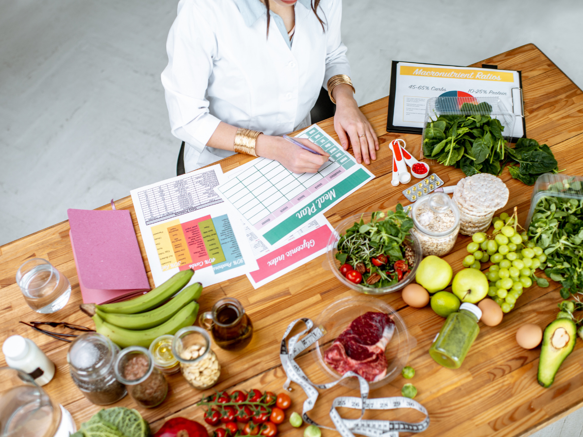 A person fills out a meal plan at a table with fresh fruits, vegetables, grains, meats, meal planning sheets, a tape measure, and nutritional guides—perfect for organizing balanced options or exploring vegetarian catering ideas.