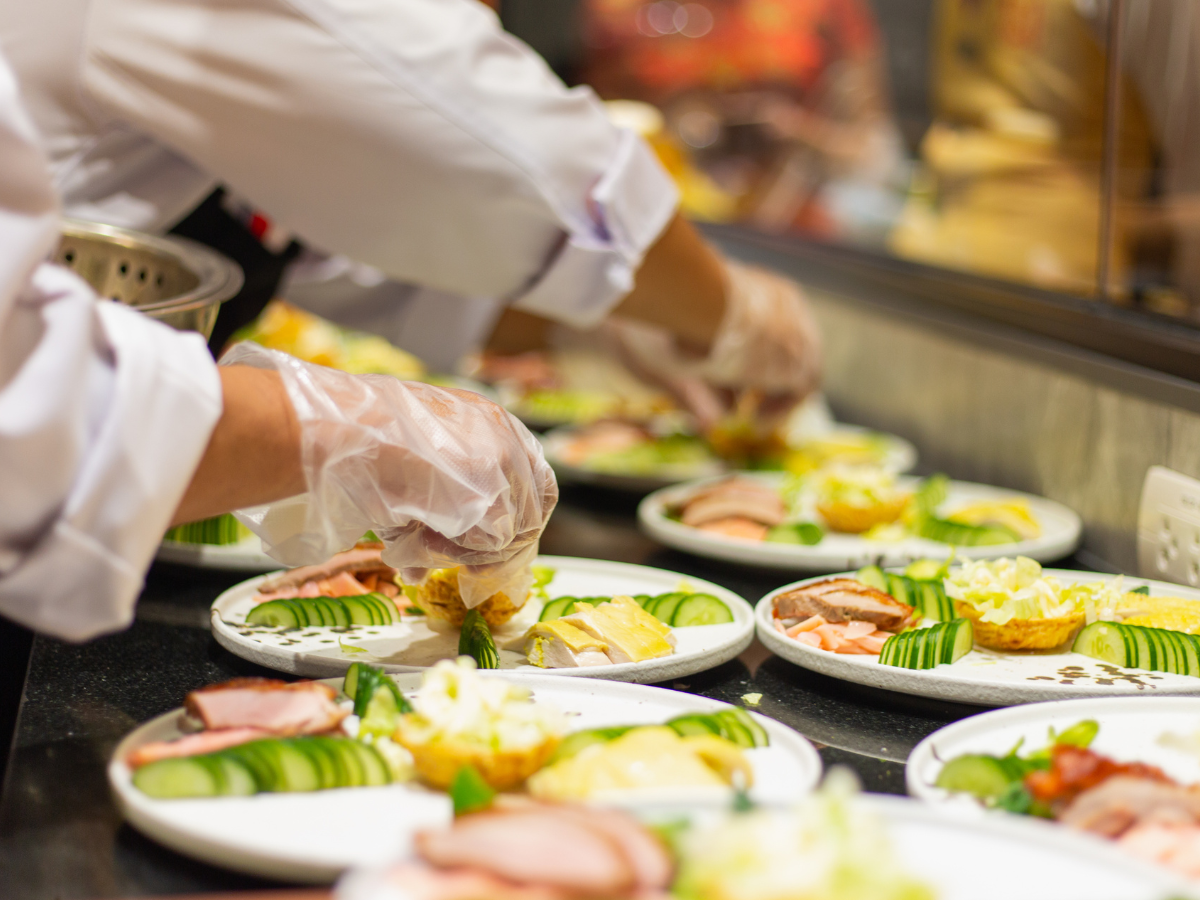 Chef wearing gloves arranges assorted food on several white plates, including sliced cucumbers, meats, and bread, in a kitchen or buffet setting featuring custom catering menus.