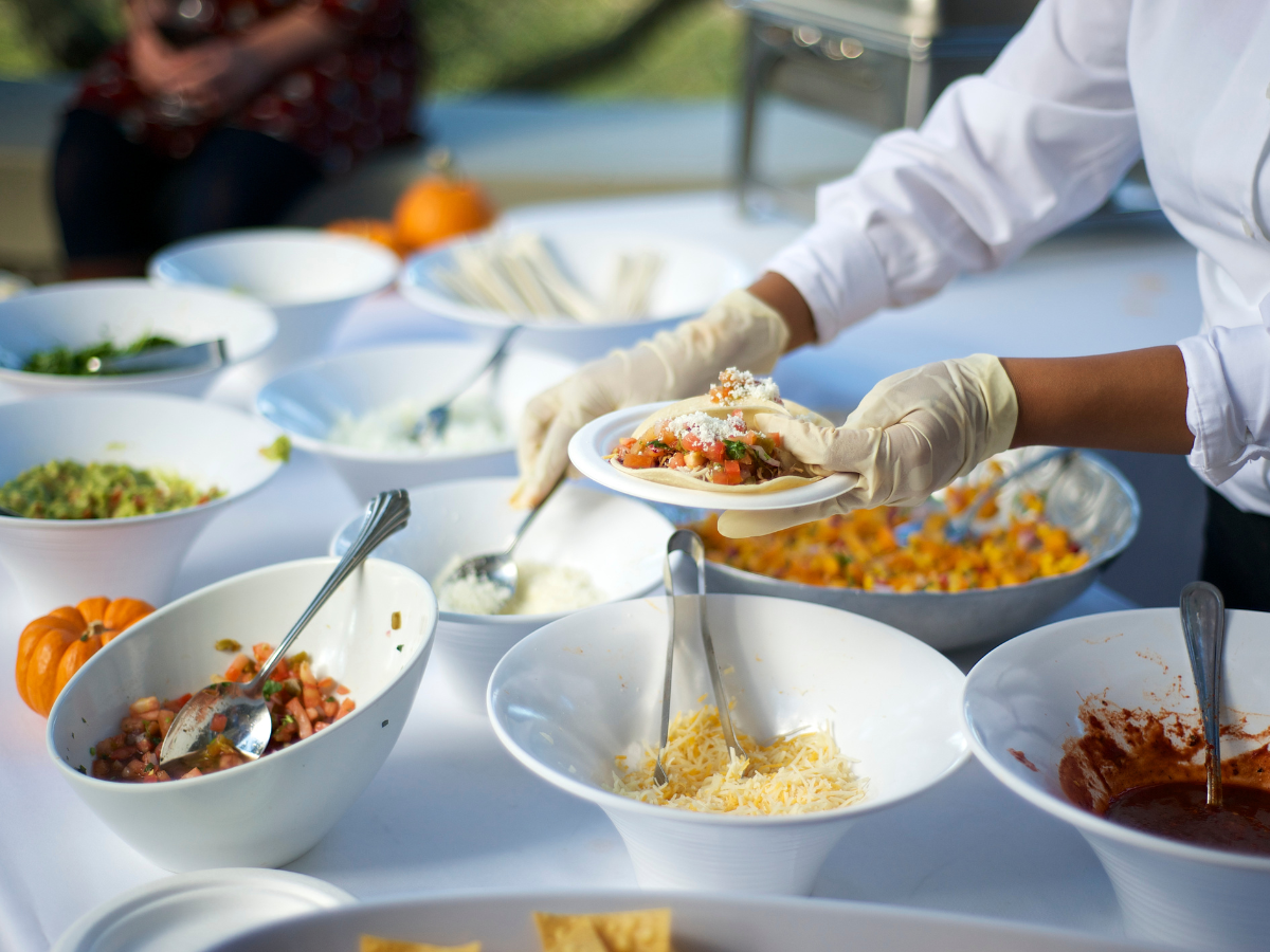 A person wearing gloves prepares a plate of food at a buffet table with various bowls of toppings and ingredients, showcasing the versatility of custom catering menus. Small pumpkins are visible on the table.