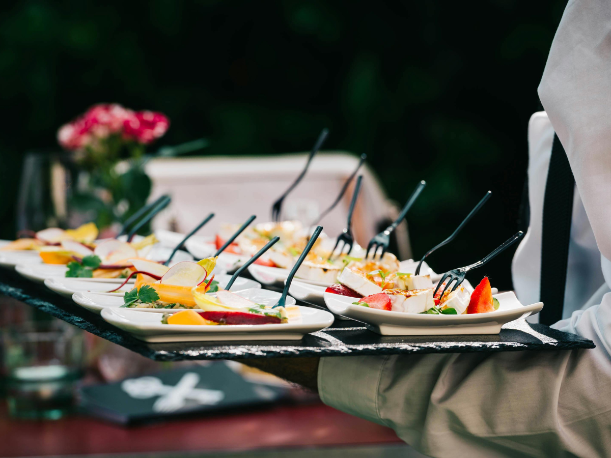 A server carries a tray with small plates of assorted appetizers, each with a black fork—a visual delight that hints at the possibilities of custom catering menus. The background is blurred, and a vase of flowers is visible.
