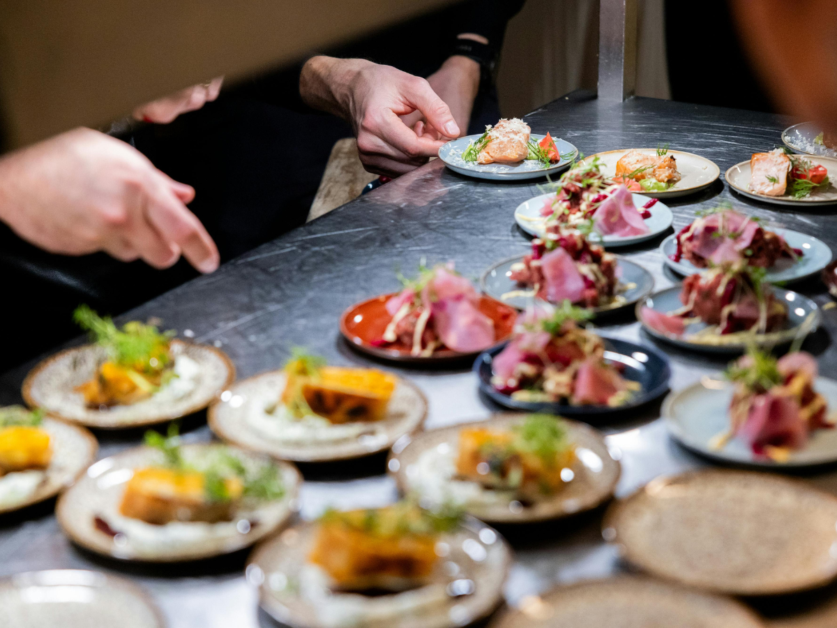 Several small plates with assorted appetizers and garnishes are arranged on a stainless steel counter, showcasing options from our custom catering menus, while people behind the counter prepare and serve the dishes.