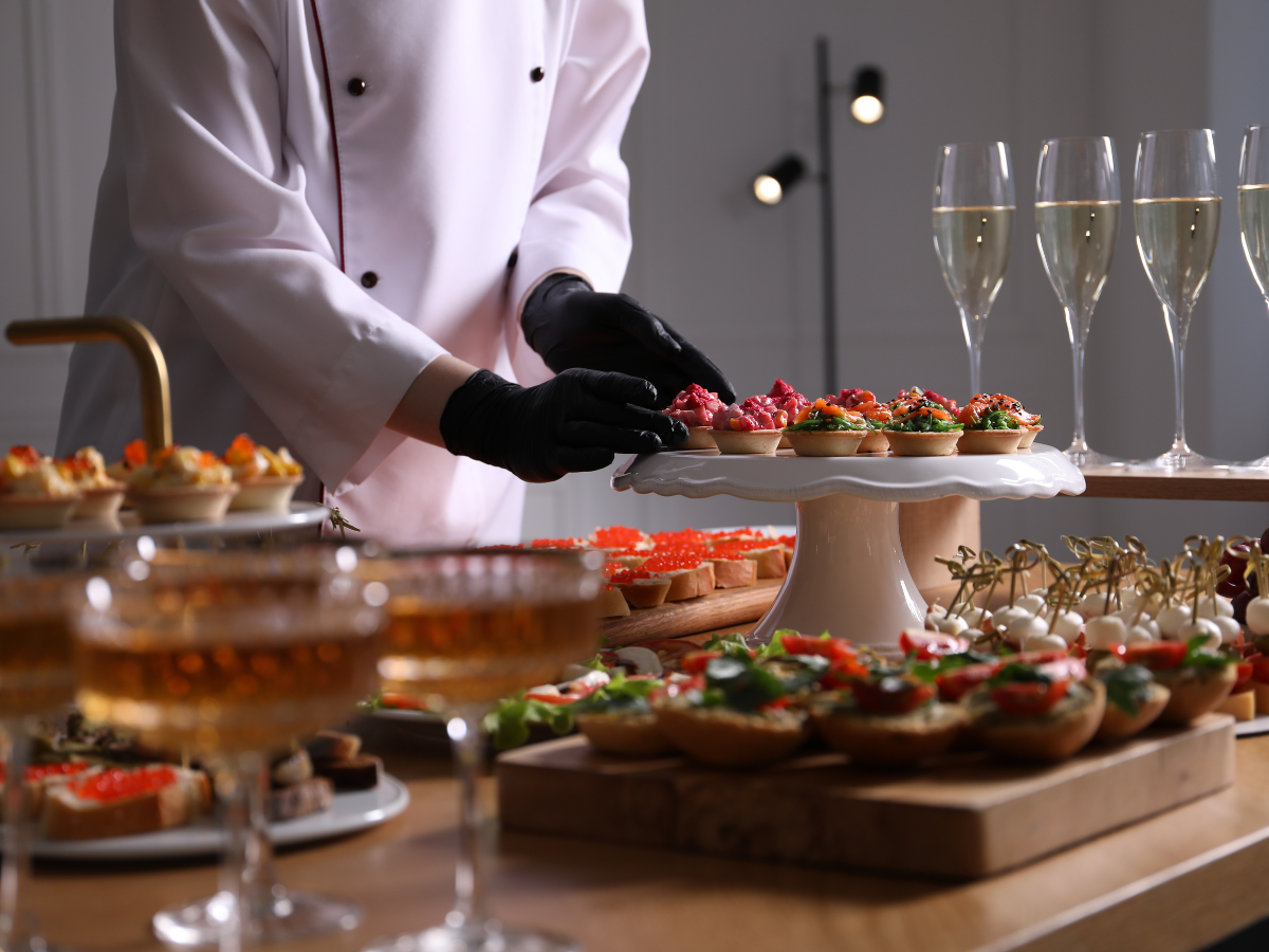 A chef in a white coat and black gloves arranges appetizers on a stand, with glasses of champagne and various finger foods on a wooden table, showcasing the artistry behind custom catering menus.