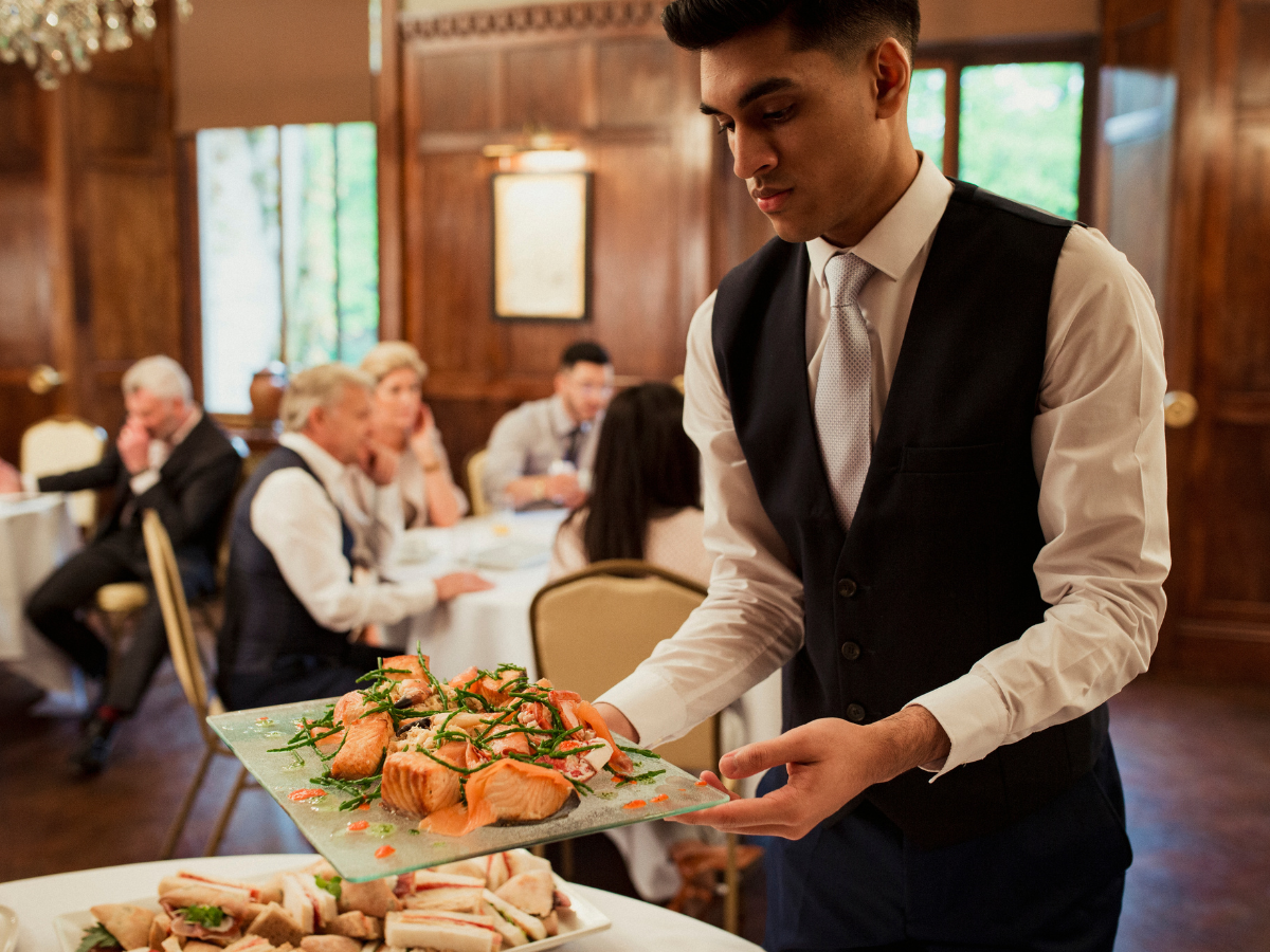 A waiter in a formal vest and tie carries a glass platter of salmon garnished with herbs for Atlanta events, serving the dining guests.
