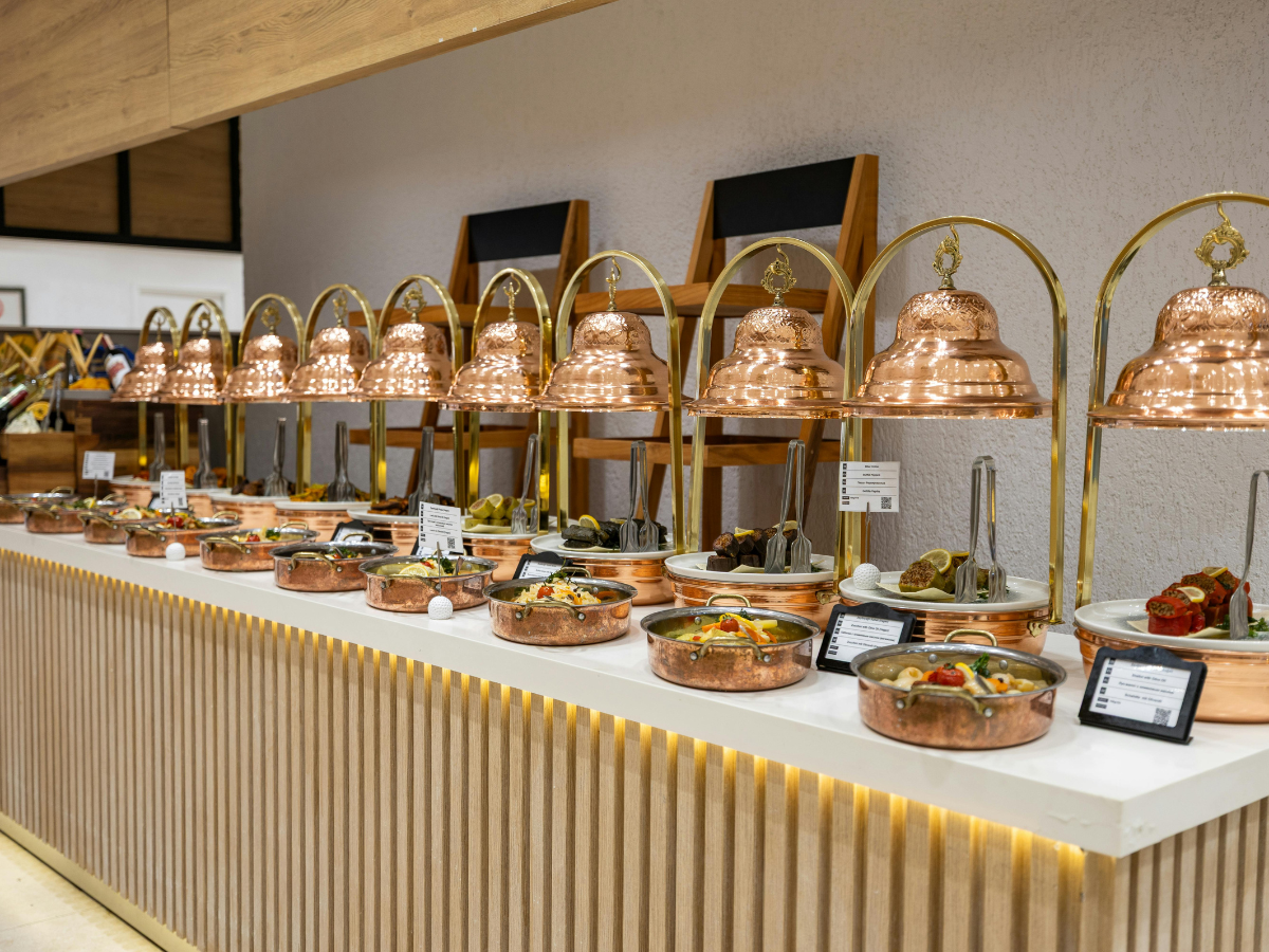 A buffet line of catering equipment rental featuring rows of copper serving dishes and matching domed lids, each labeled with a small sign.