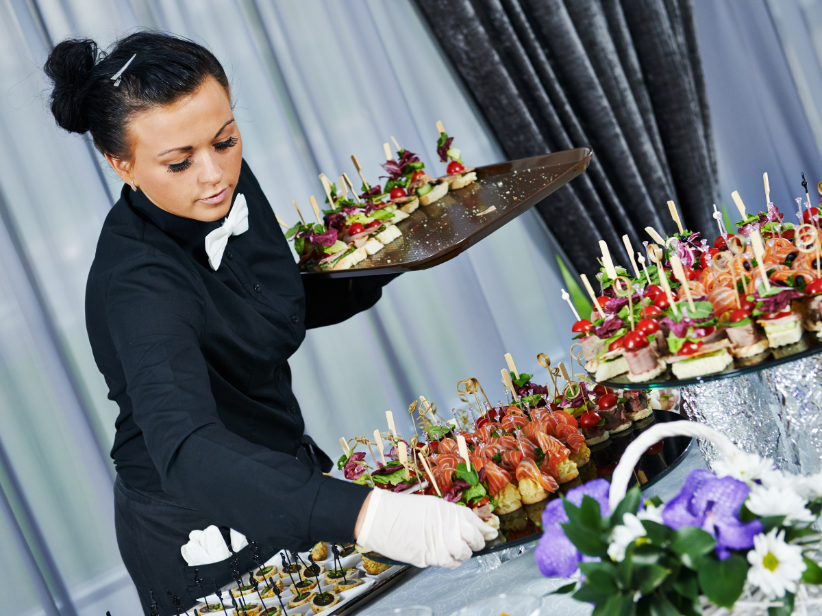 A server in a black uniform and white gloves arranges trays of assorted appetizers using Atlanta event rentals at a buffet.
