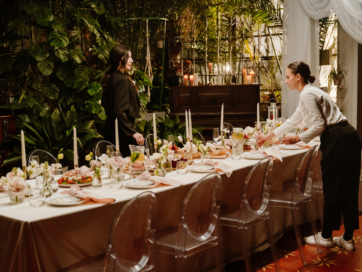 Two women set a long dining table with catering equipment rental in a room filled with potted plants.