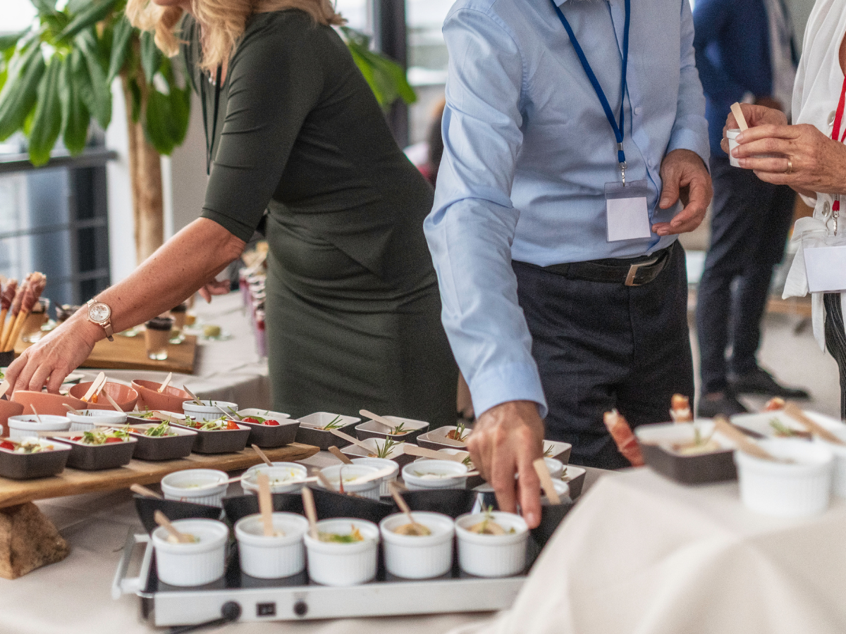 People serve themselves food from a buffet table with small dishes and ramekins at a corporate conference catering event; only their torsos and arms are visible.