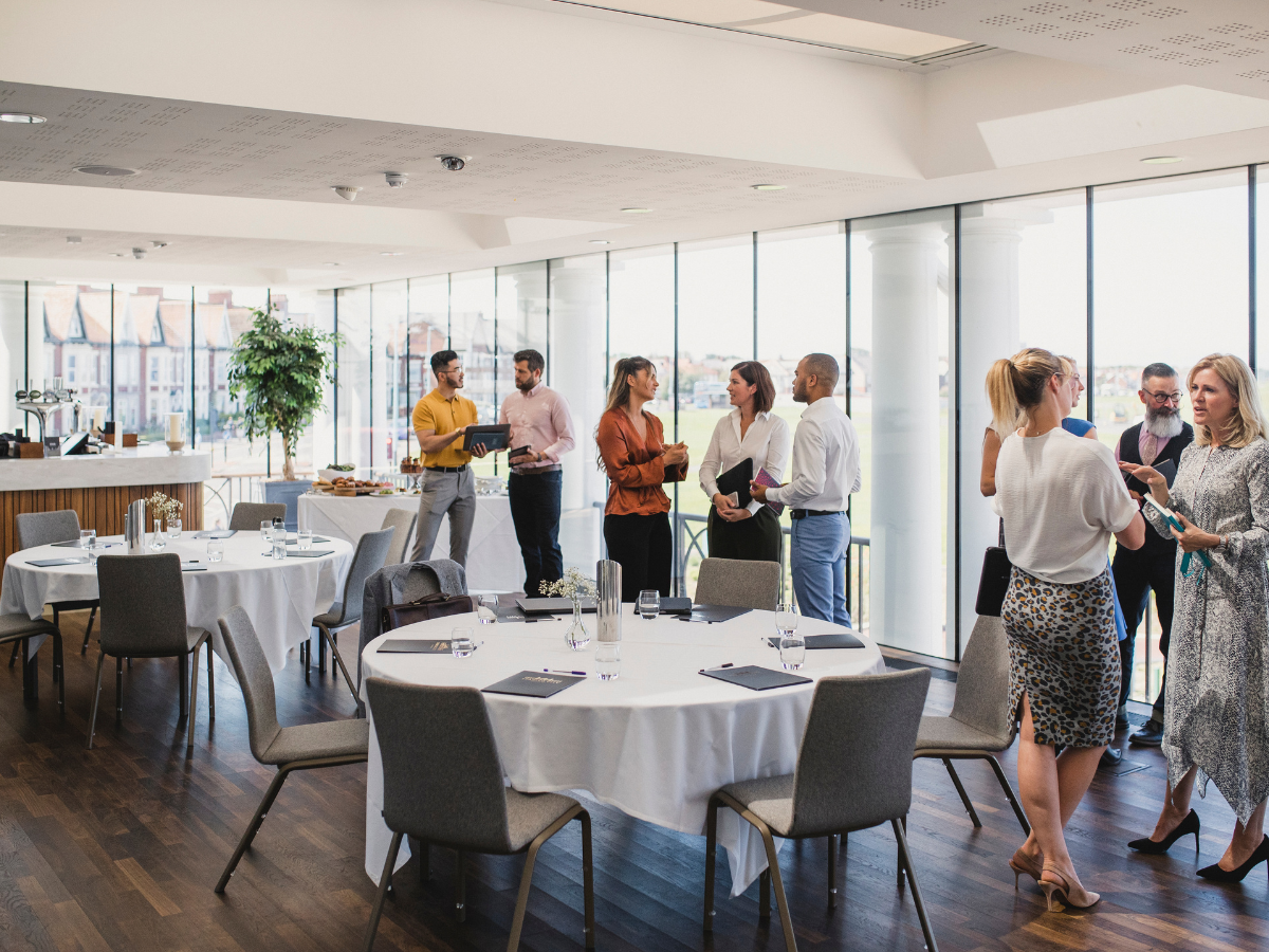 People stand and converse in a bright, modern conference room with round tables, corporate conference catering, notepads, and refreshments visible by large windows.