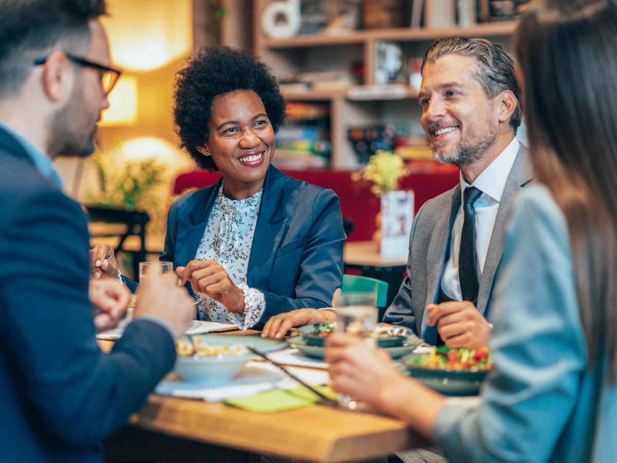 Four people in business attire sit together at a restaurant table, enjoying a meal and lively conversation, showcasing a relaxed corporate conference catering experience.