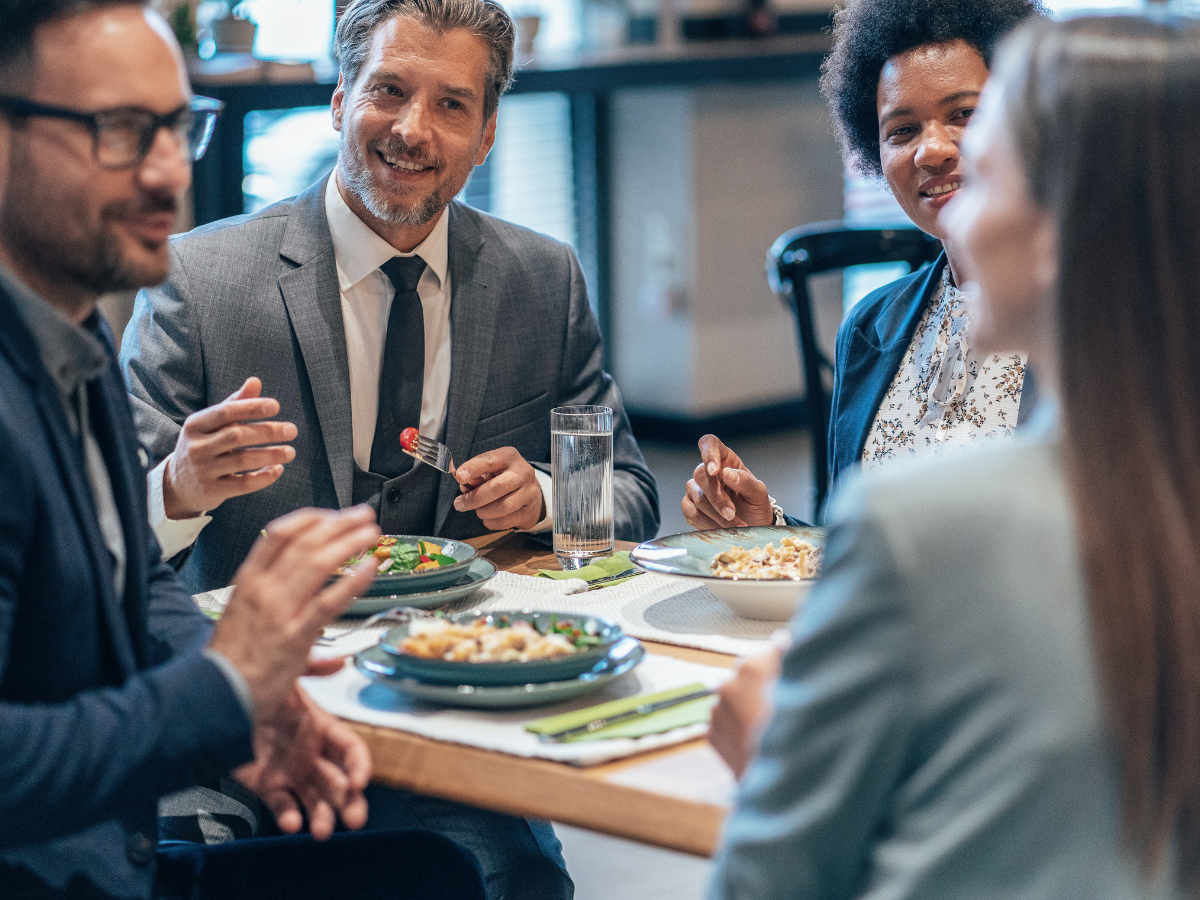 Four people in business attire sit at a table, enjoying a meal together with corporate conference catering in a restaurant or caf&eacute; setting.
